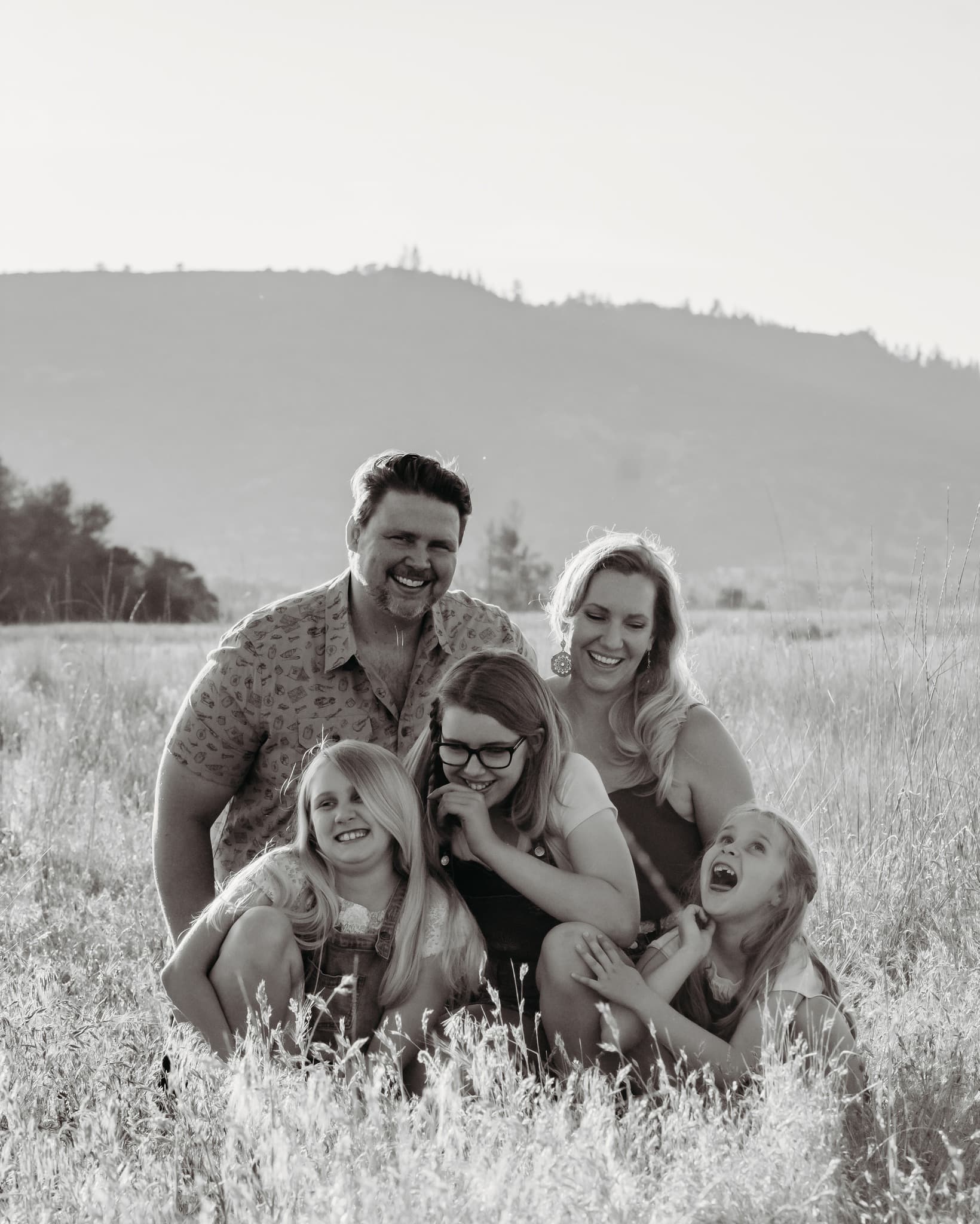 Laughing family of five huddles together in a grassy field with hills in the background.